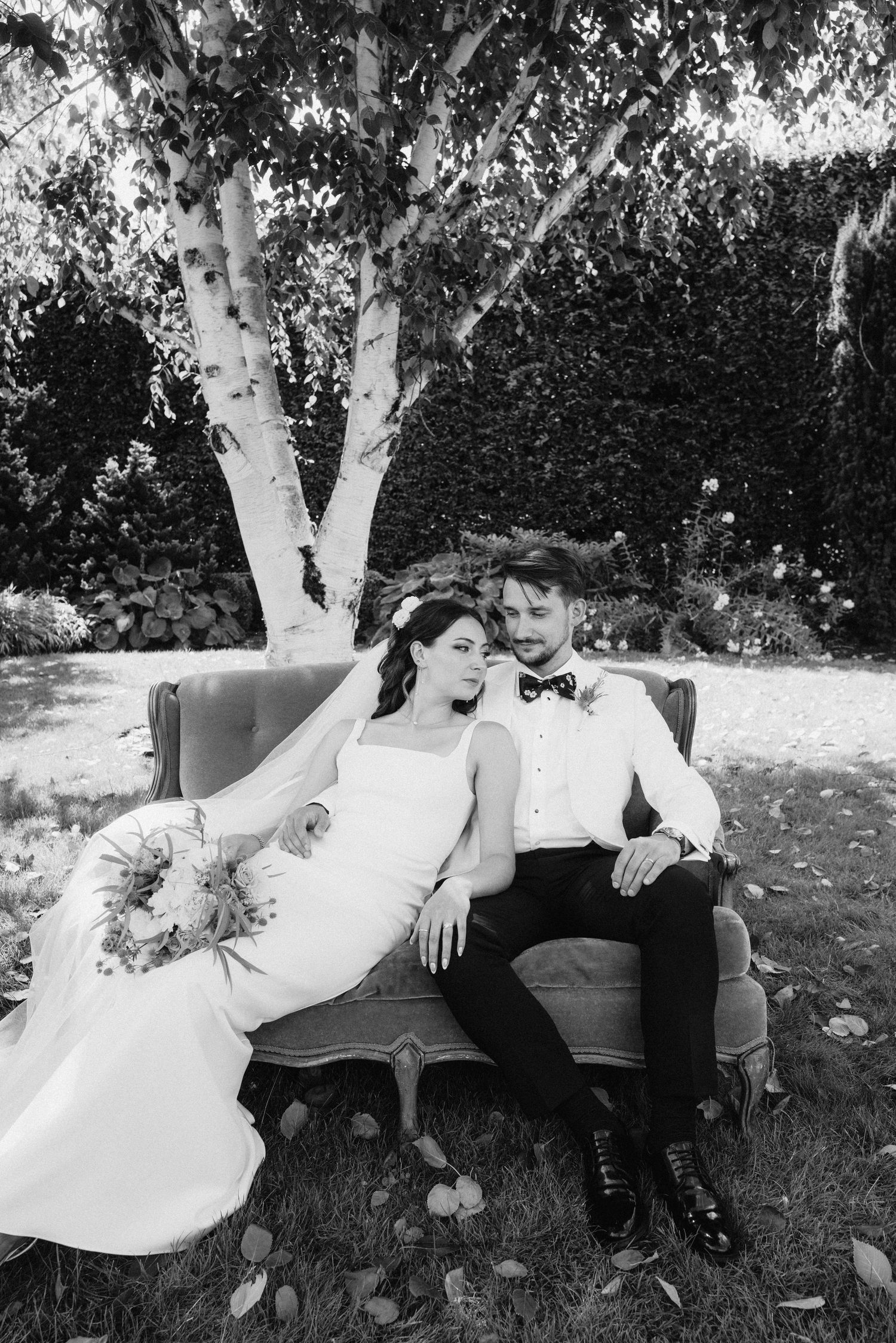 Wedding couple relaxes on vintage brown couch under birch trees in garden setting.