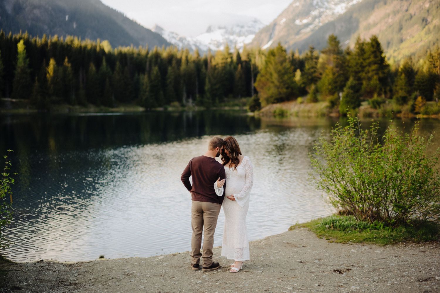 A couple shares a special moment on a rocky beach with mountains and forest reflecting in the lake.