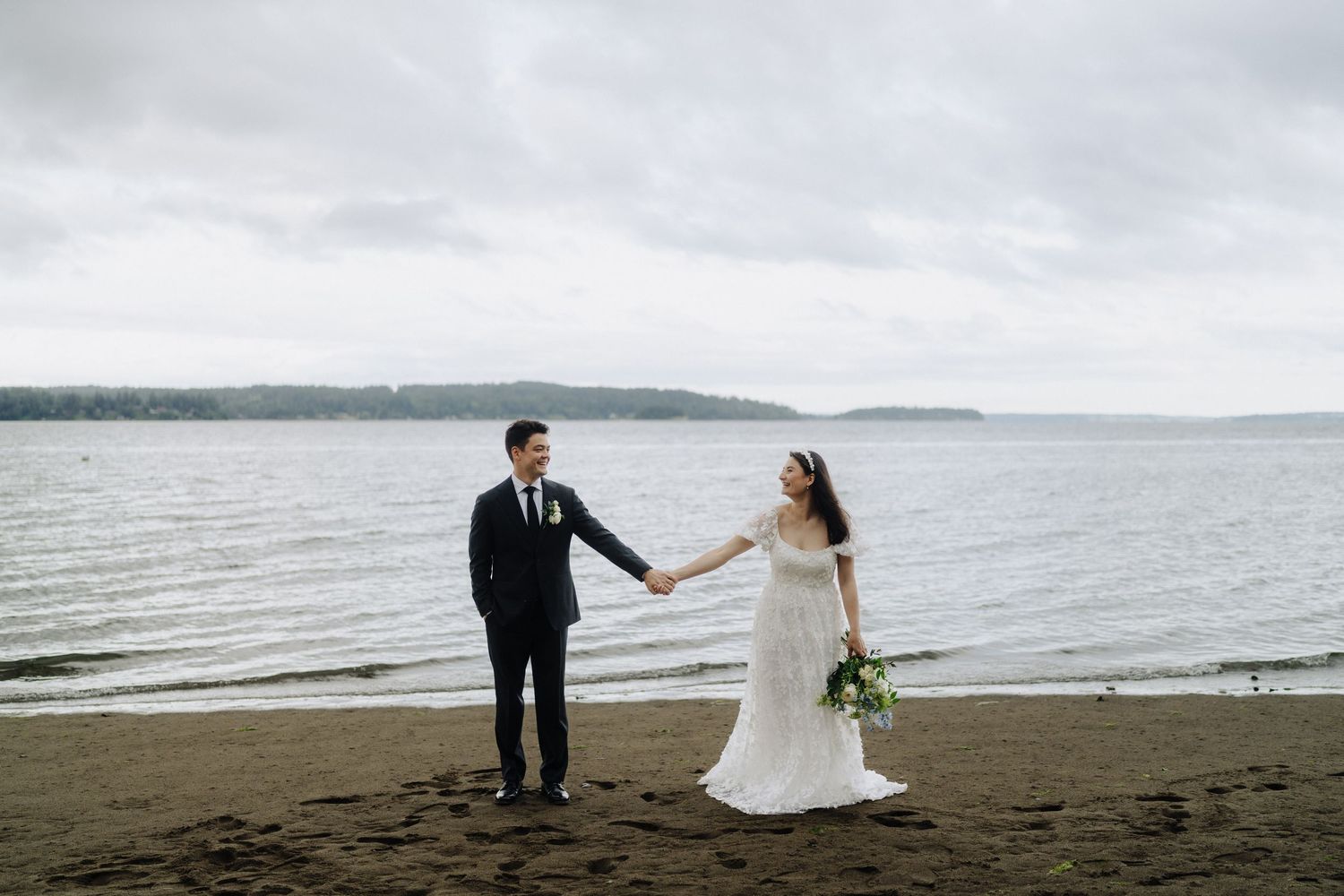Newlyweds walk along a misty beach shoreline with dramatic cloudy sky.