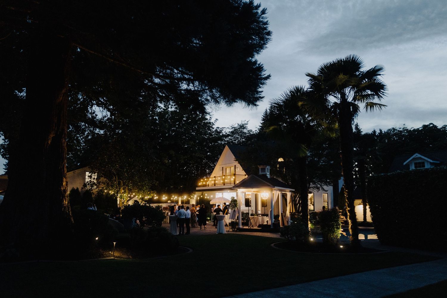 Panoramic twilight view of an illuminated wedding venue with string lights and glowing windows.