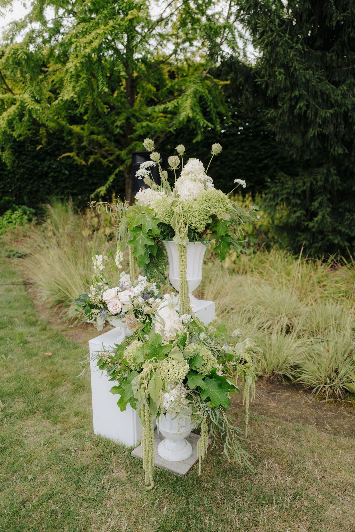 White floral arrangements in elegant urns displayed on pedestals against a green garden backdrop.