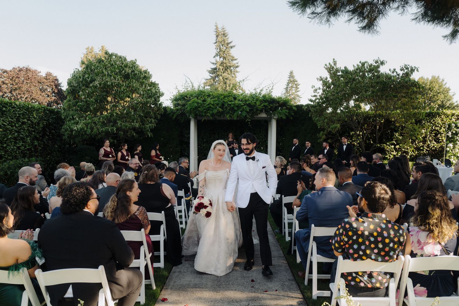 Wedding guests seated in white chairs watch an outdoor ceremony as sunlight filters through trees.