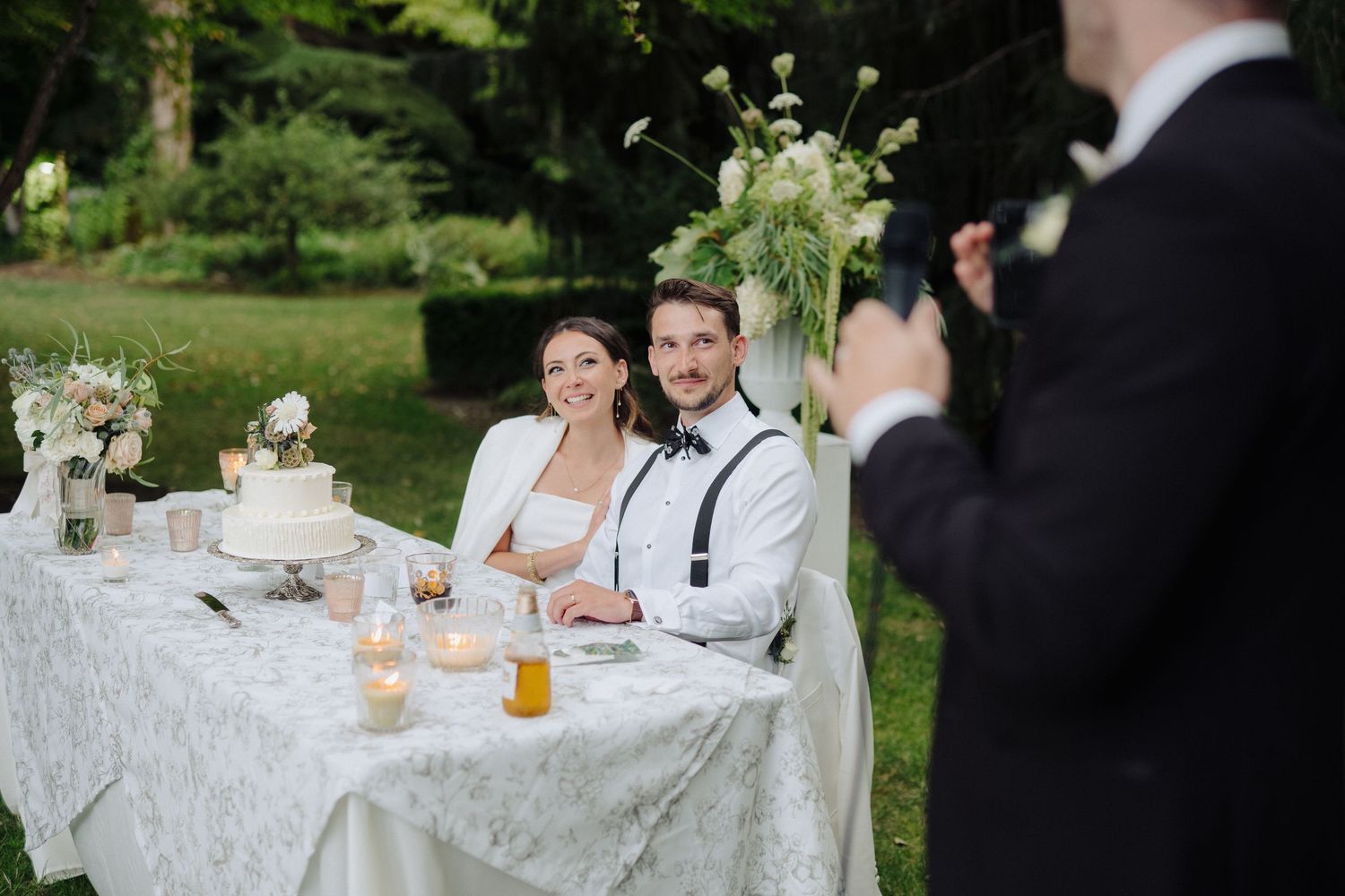 Wedding reception guests seated at elegant white-clothed tables in a garden setting.