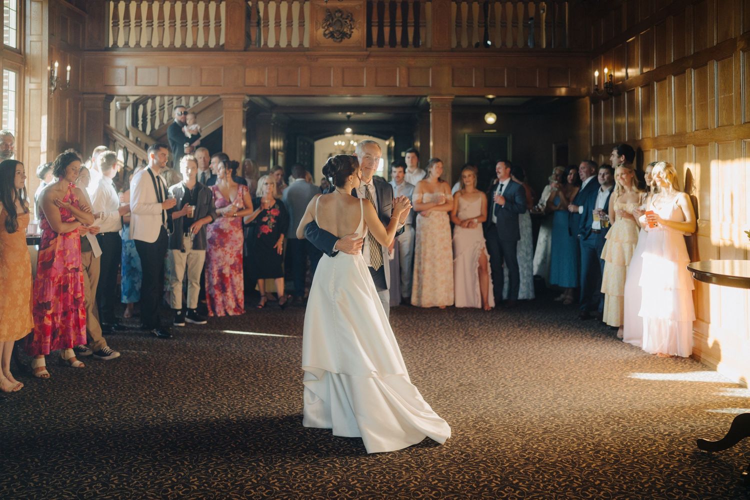 Wedding reception dance in grand wooden hall with guests watching from the sides.