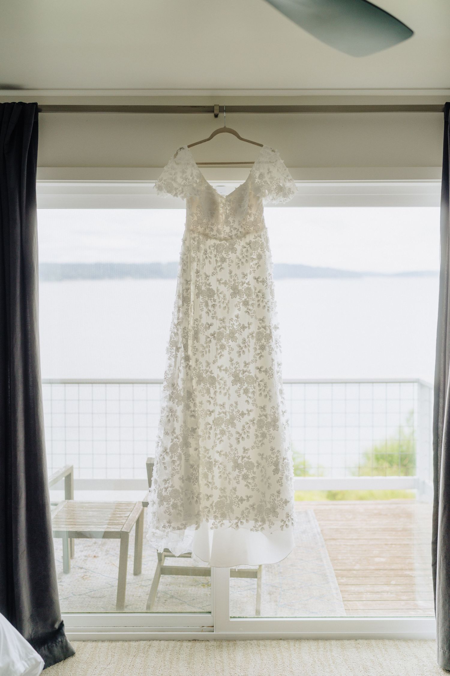 A delicate white lace wedding dress hangs by a window overlooking a scenic waterfront view.