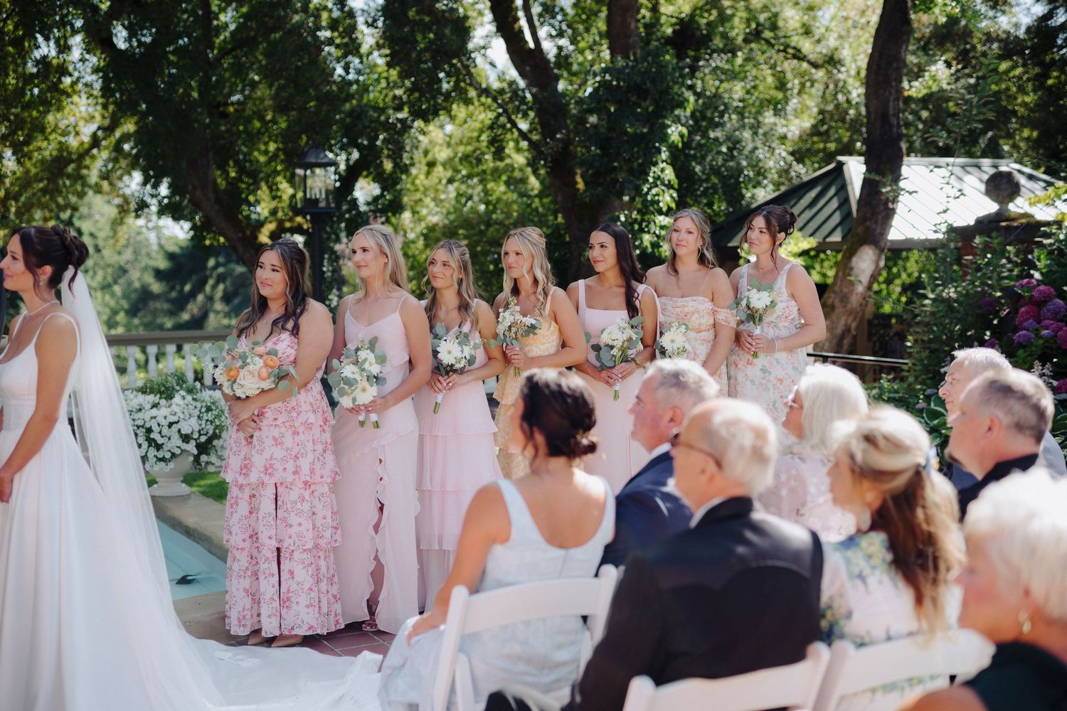 Wedding guests gather under trees for an outdoor ceremony with bridesmaids in pink dresses.