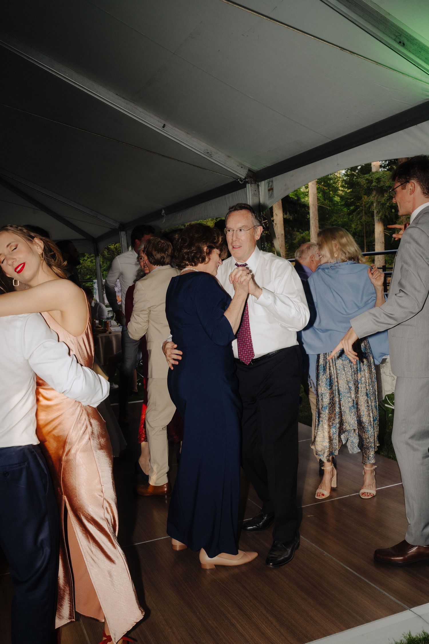 Wedding reception guests dancing under a white tent on a wooden dance floor during evening celebrations.