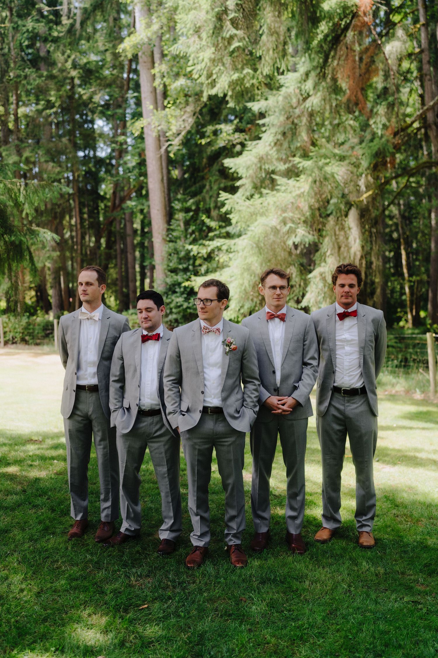 Groomsmen in matching gray suits and red bowties line up against a backdrop of evergreen trees.