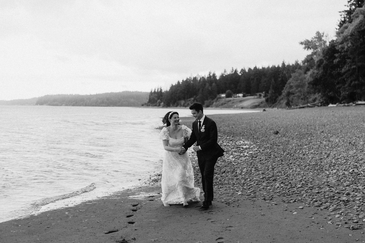 A scenic beach wedding photoshoot with evergreen trees and rocky shoreline in the background.