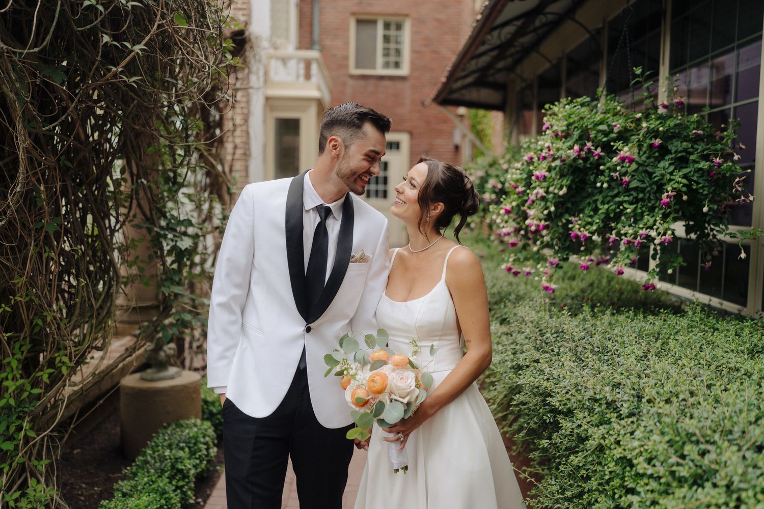Wedding party poses in front of a brick building surrounded by lush greenery.