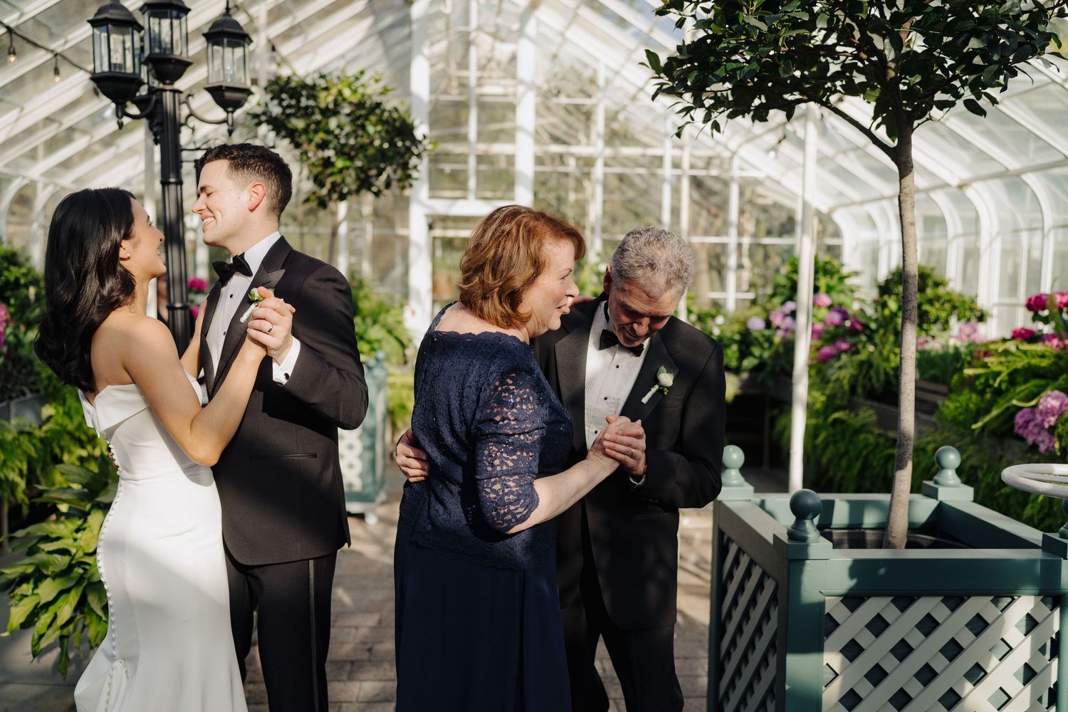 Two couples share a dance moment in an elegant greenhouse setting with hanging lanterns.