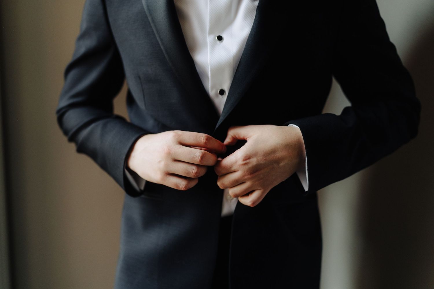Close-up shot of hands buttoning a black formal suit jacket with white dress shirt visible underneath.