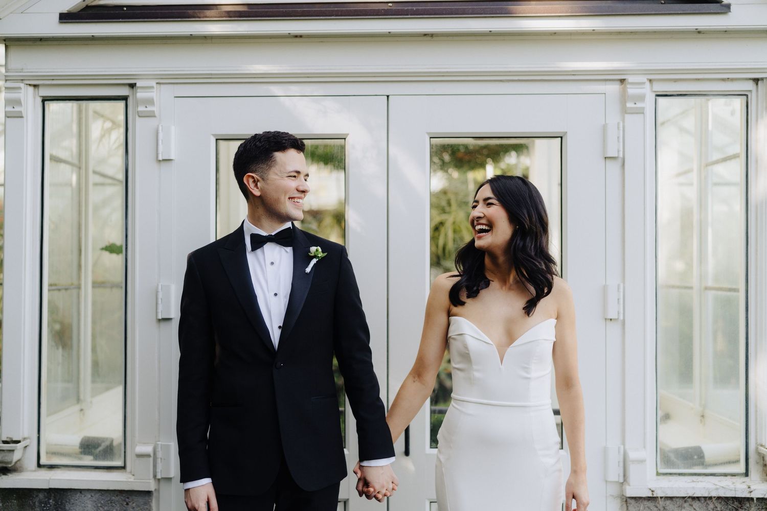 Newlyweds in formal wedding attire smiling and holding hands outside a white-framed entrance.