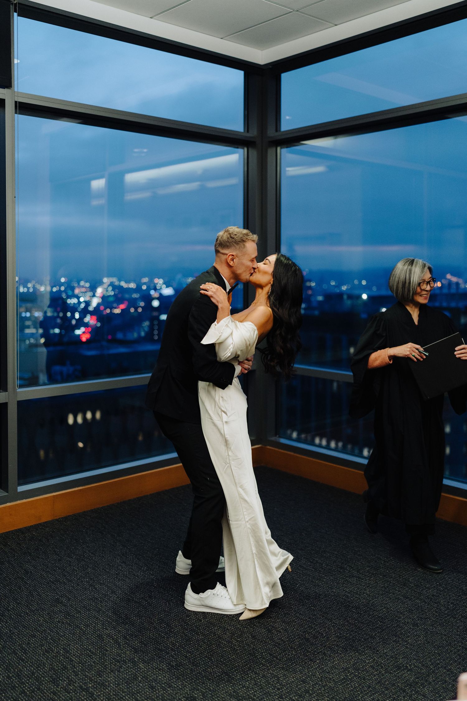 A couple in formal attire dances near floor-to-ceiling windows with a cityscape view at night.
