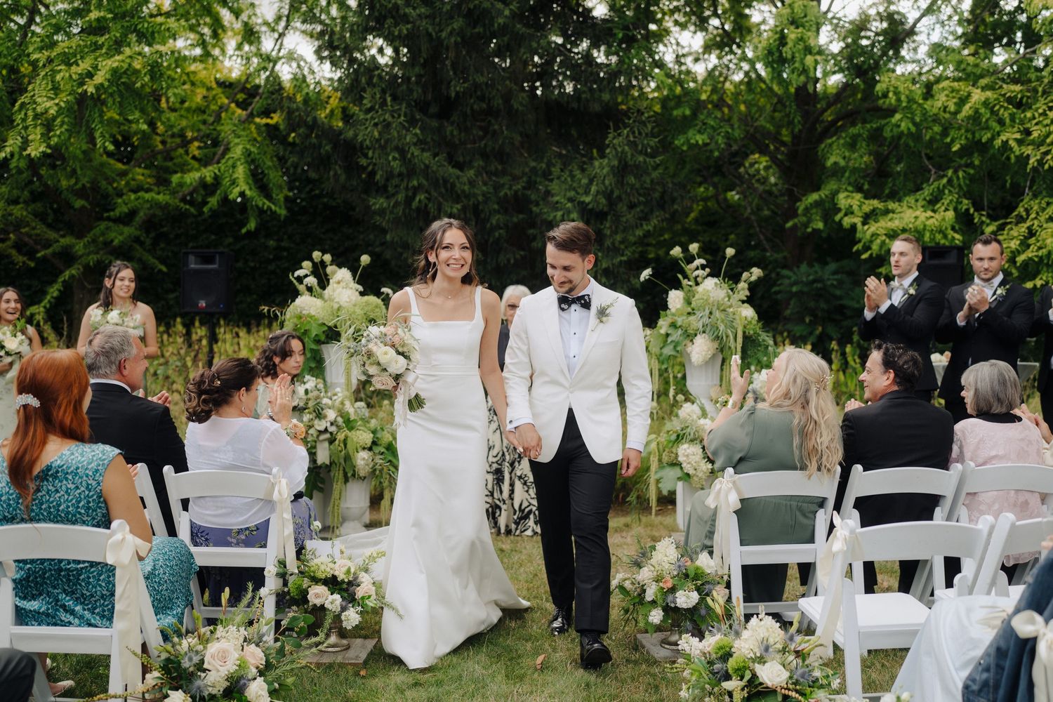 Garden wedding ceremony with guests seated in white chairs surrounded by greenery.