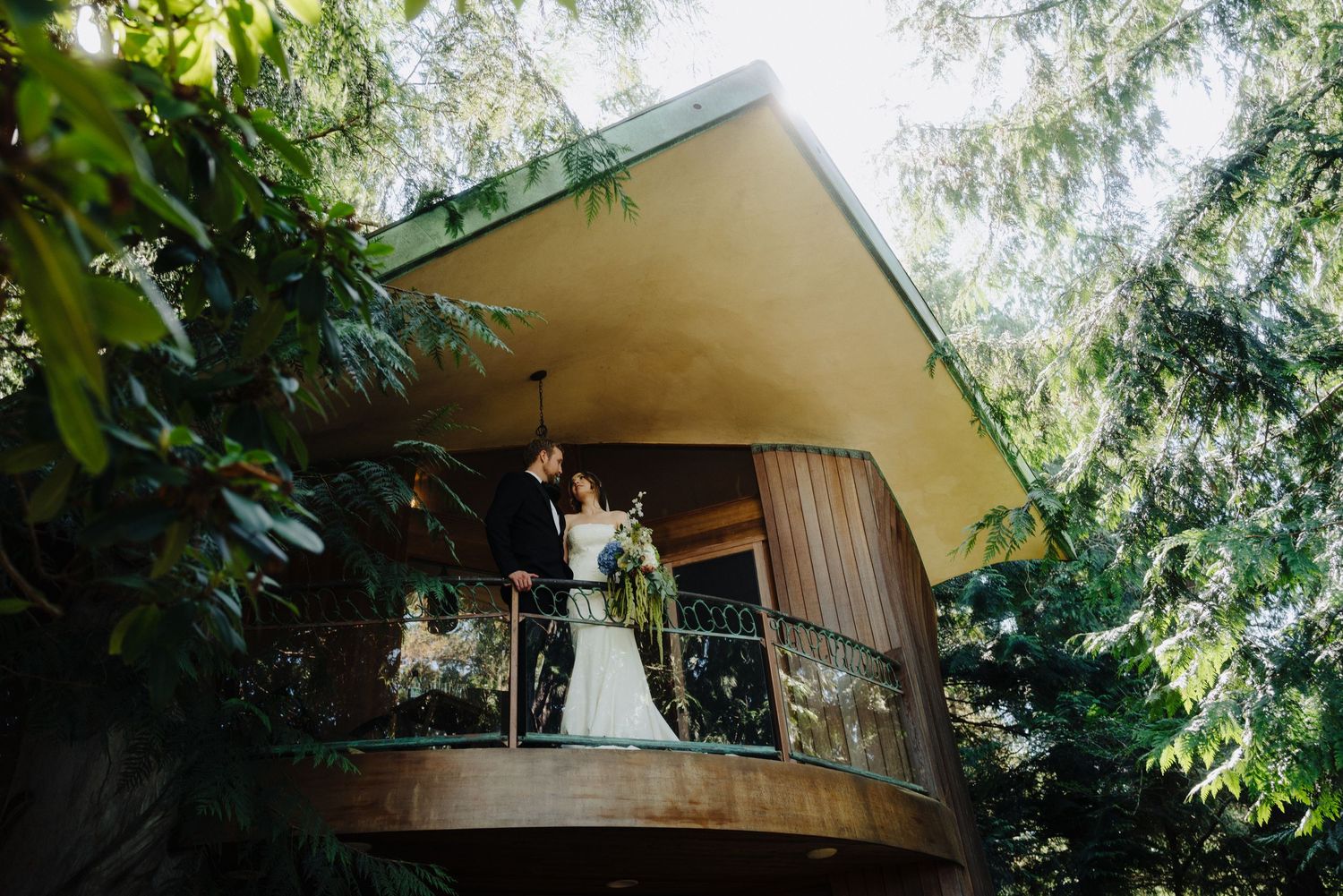 Newlyweds pose on the balcony of a unique A-frame structure surrounded by tall trees.