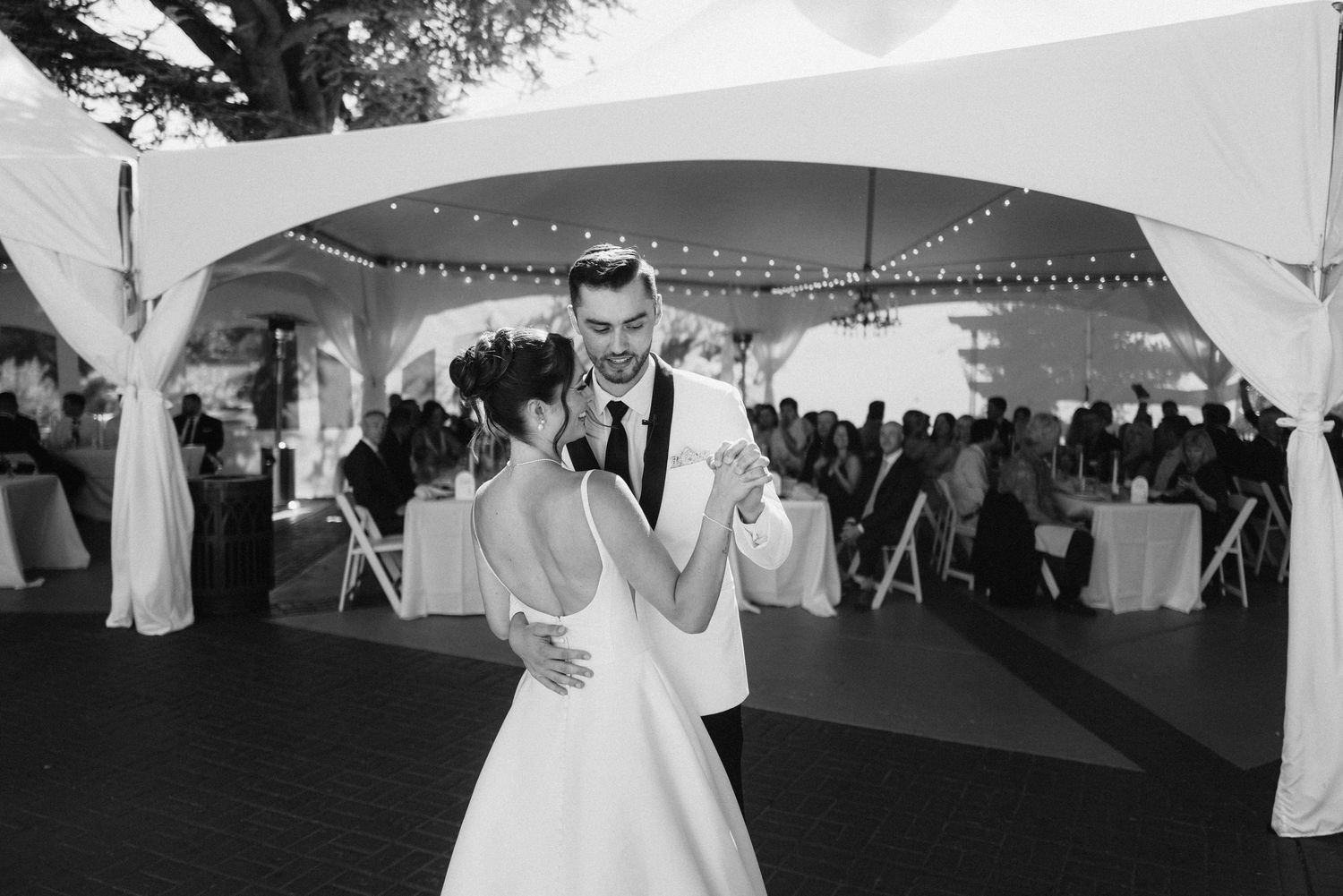 Black and white photo of a couple sharing their first dance under a white tent with string lights.