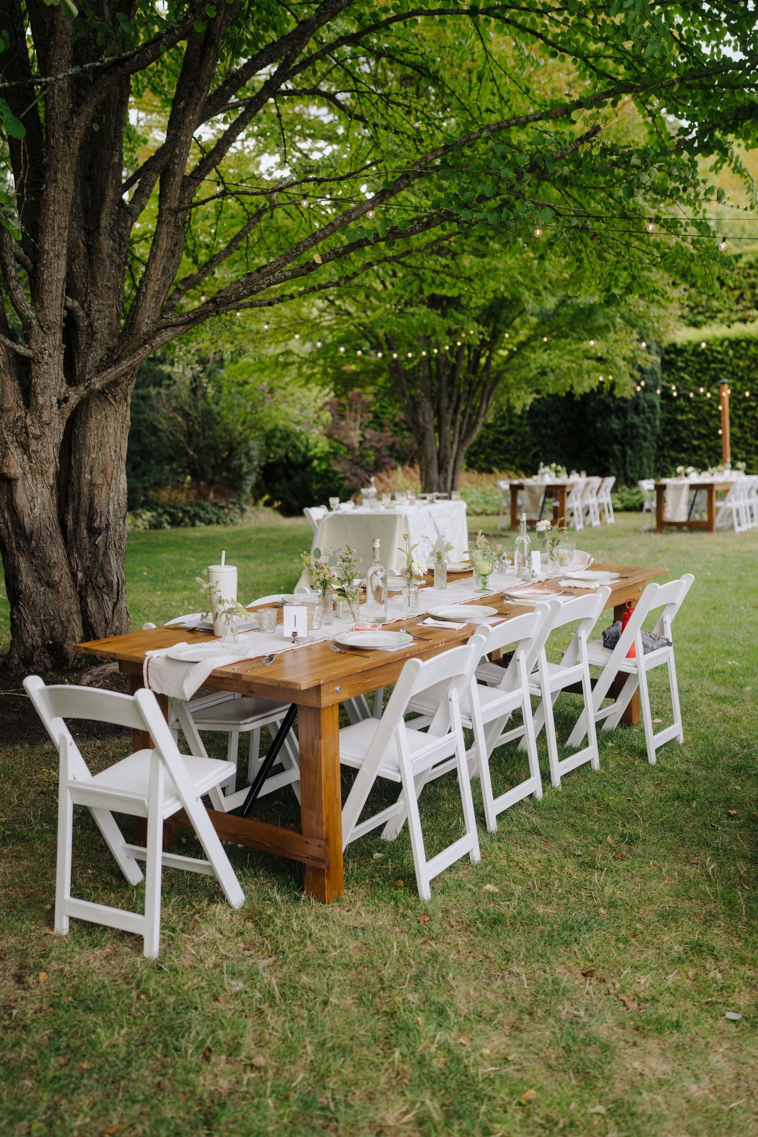 Long wooden farm tables with white chairs set up outdoors under mature trees for an elegant garden wedding reception.
