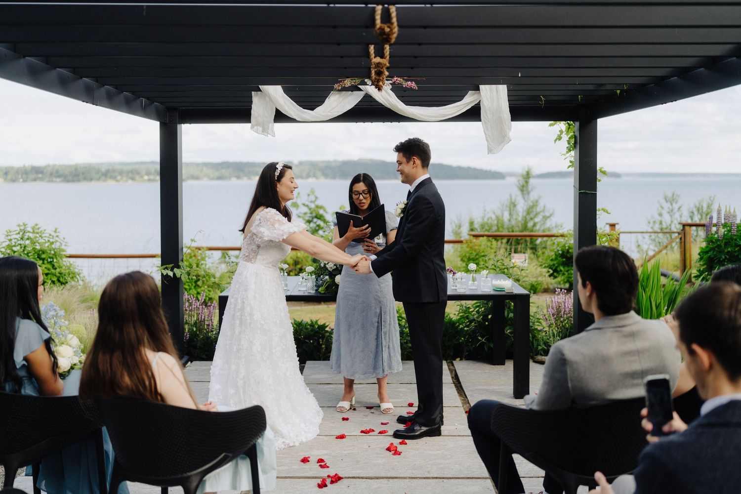 Wedding ceremony setup with guests seated under a covered outdoor pavilion by the water.