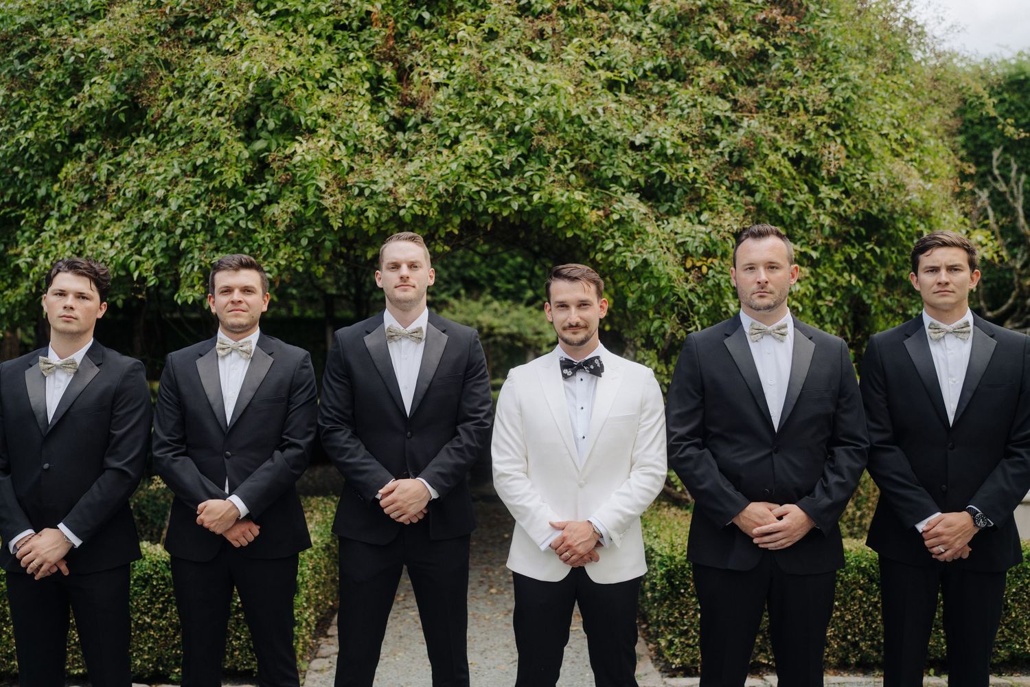 Wedding party members in formal attire lined up against a green garden backdrop.