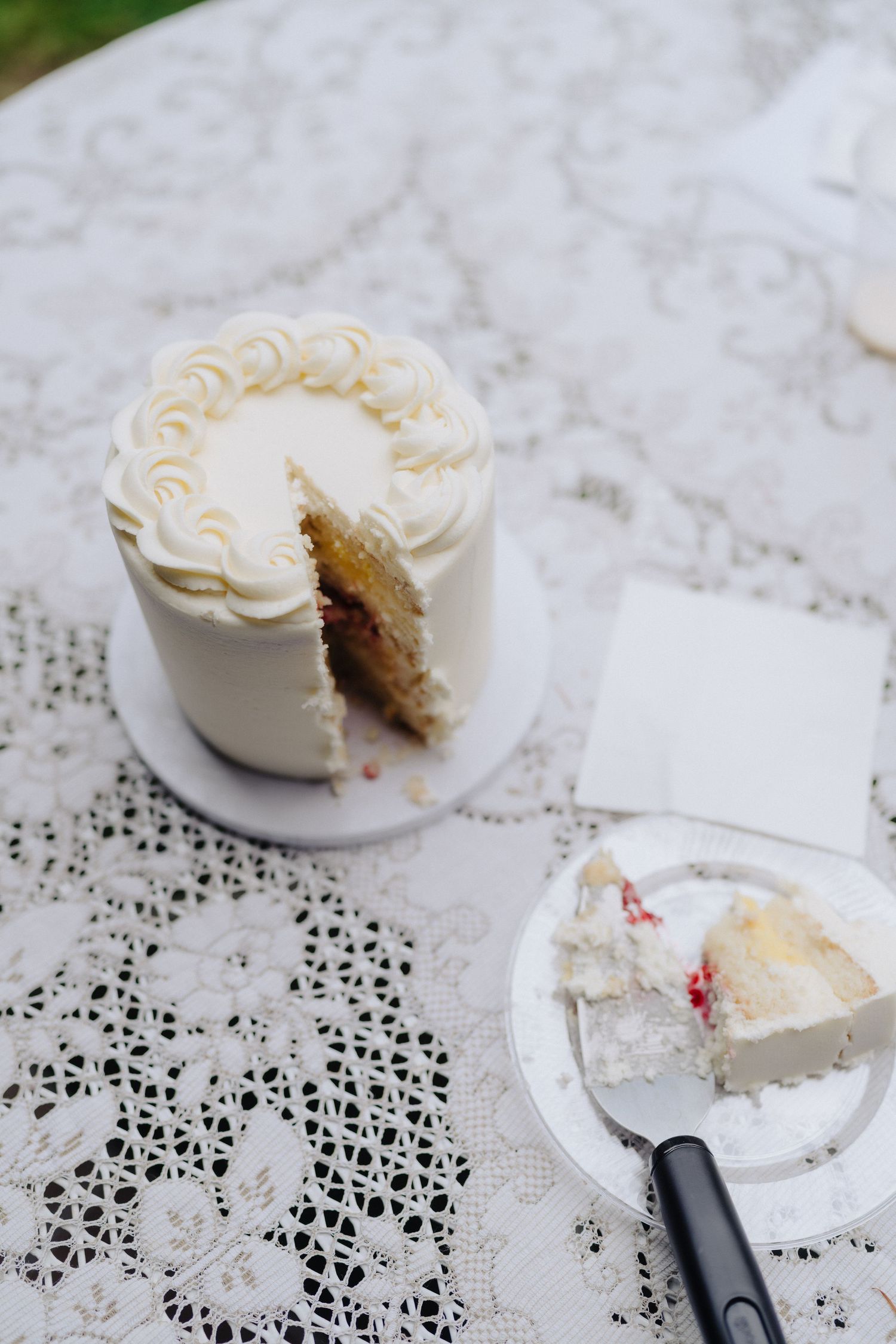 A small white wedding cake with piped frosting and a slice removed on a lace tablecloth.