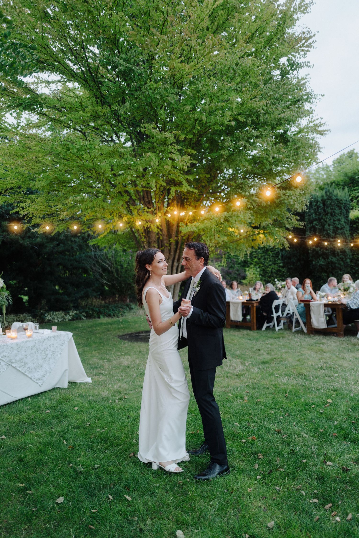 A romantic outdoor wedding dance under string lights and leafy trees.