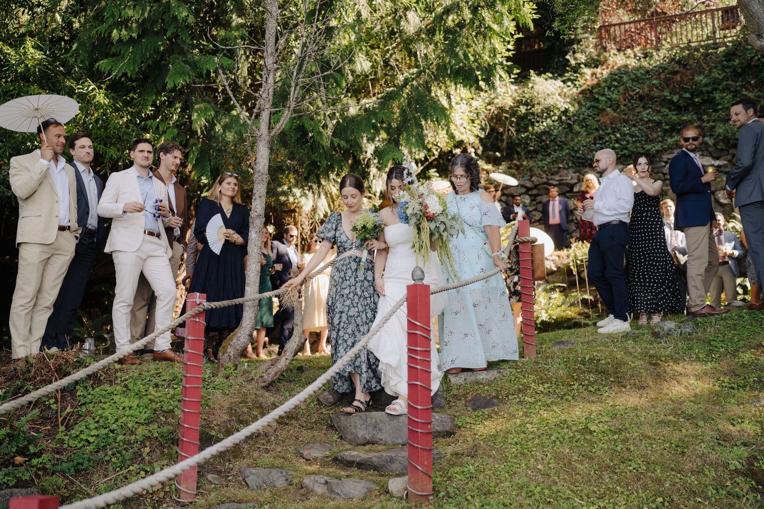 Wedding ceremony guests gather under trees in an outdoor garden setting during a summer celebration.