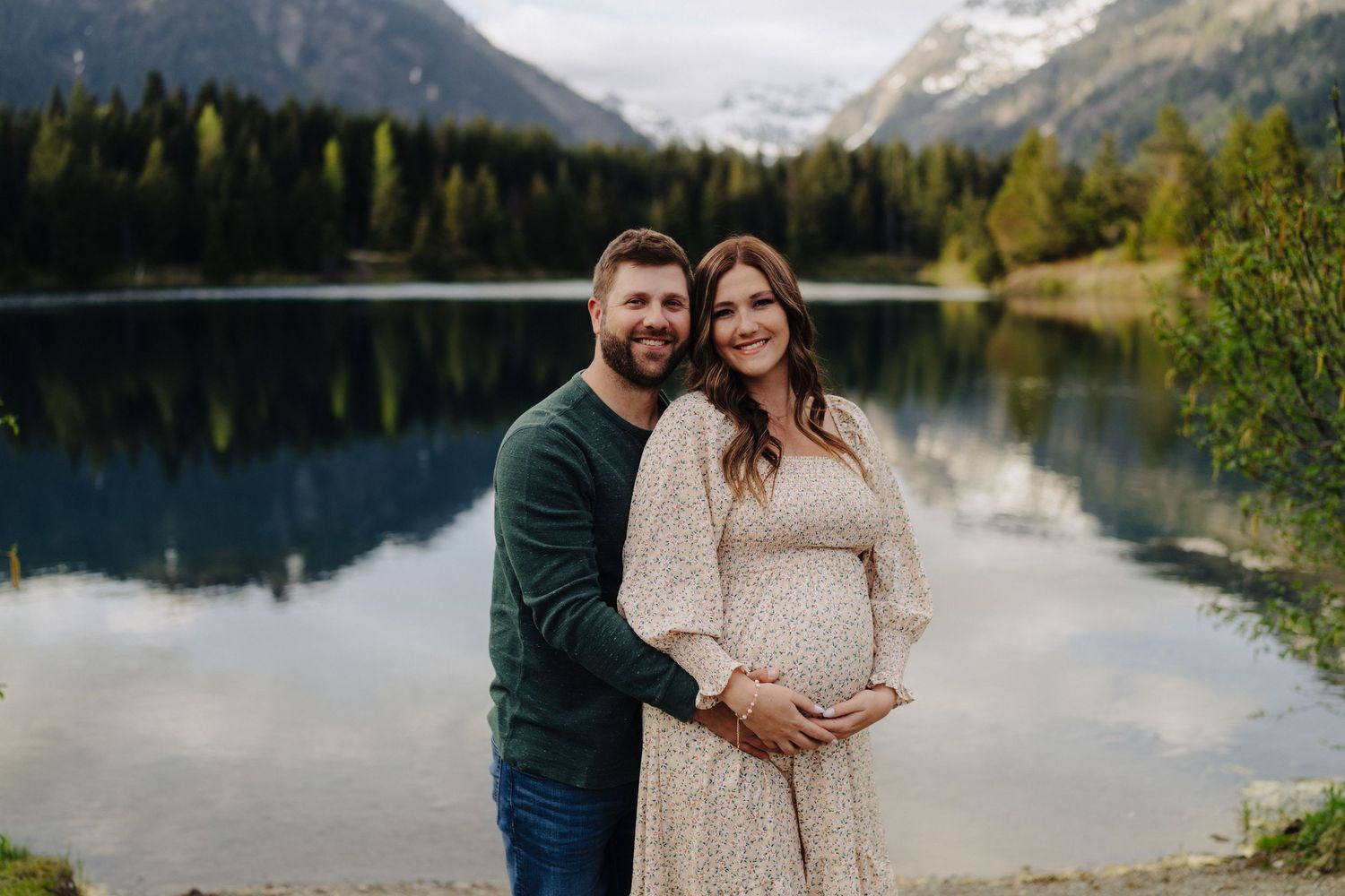 Couple embraces by a serene mountain lake with snow-capped peaks and evergreen forest in the background.