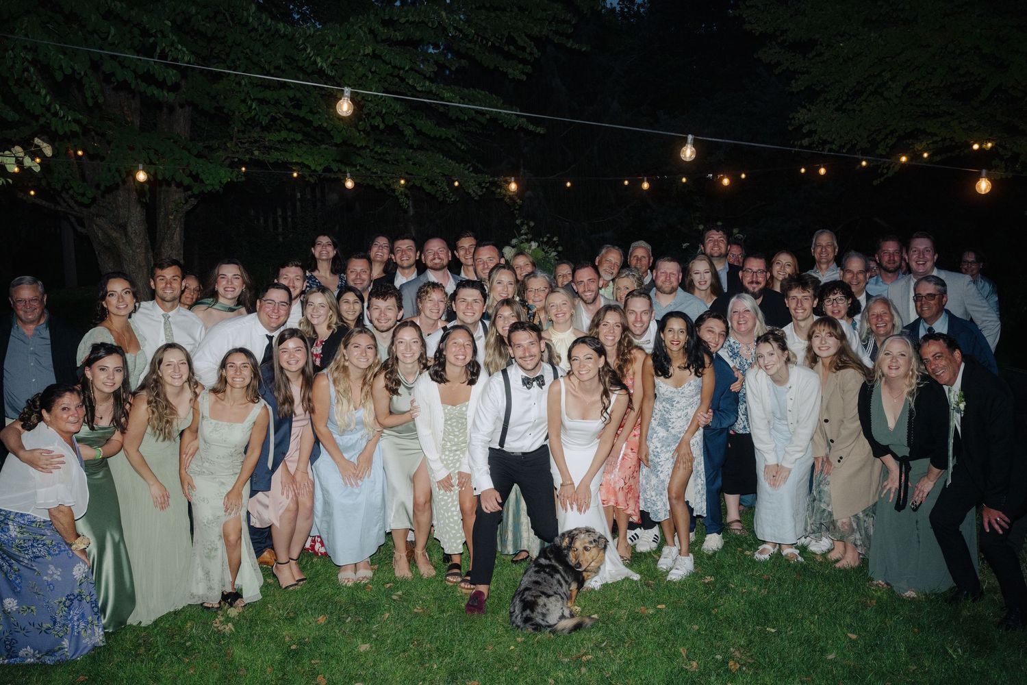 Large wedding party group photo taken outdoors at dusk with string lights overhead.