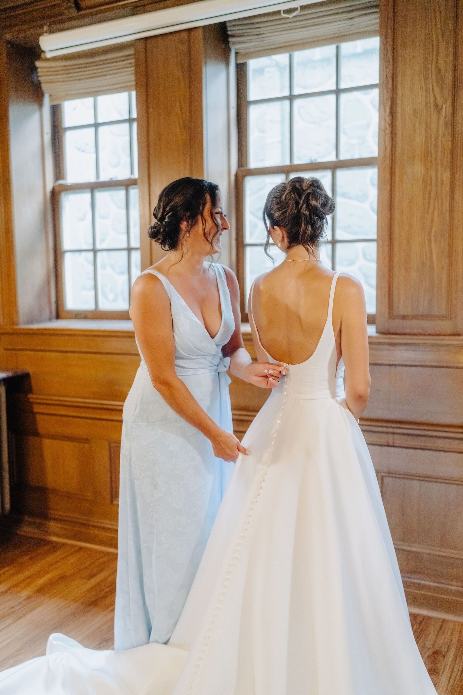 Two people in elegant dresses stand together by large windows in a wood-paneled room.