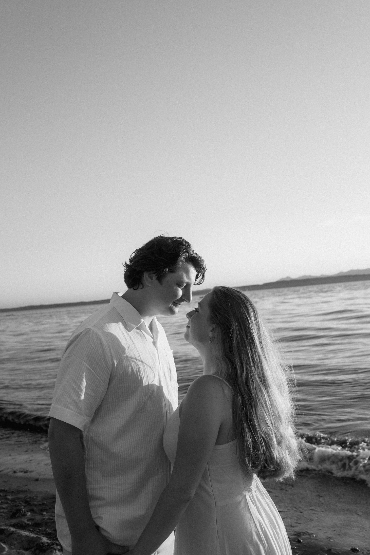 A romantic black and white photo series of a couple embracing on a beach at sunset with ocean waves in the background.