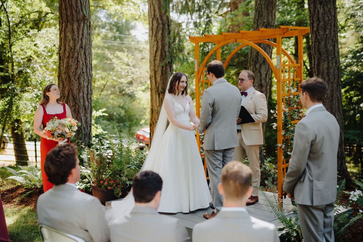 Outdoor wedding ceremony under an orange wooden arch surrounded by tall evergreen trees.