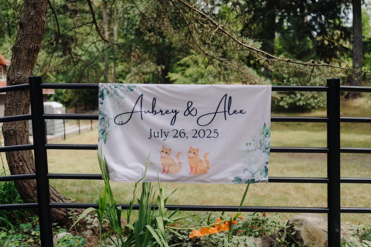 A wedding banner with floral design hangs on a rustic fence displaying names and date.