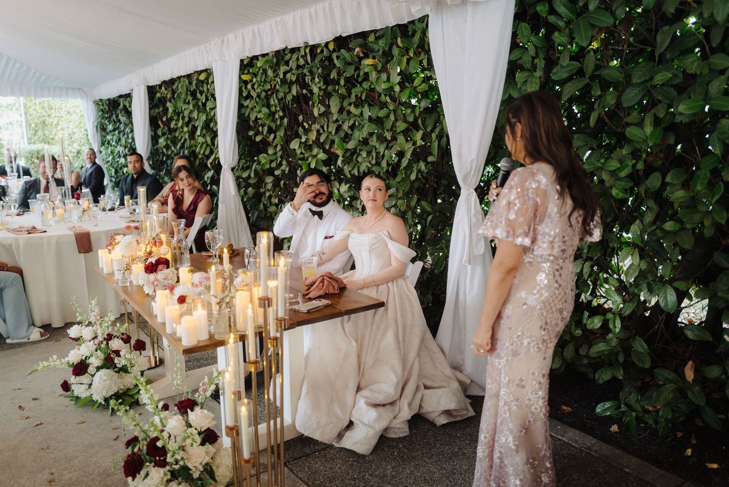 Wedding reception space decorated with hanging lanterns, white drapes and floral arrangements in a garden setting.