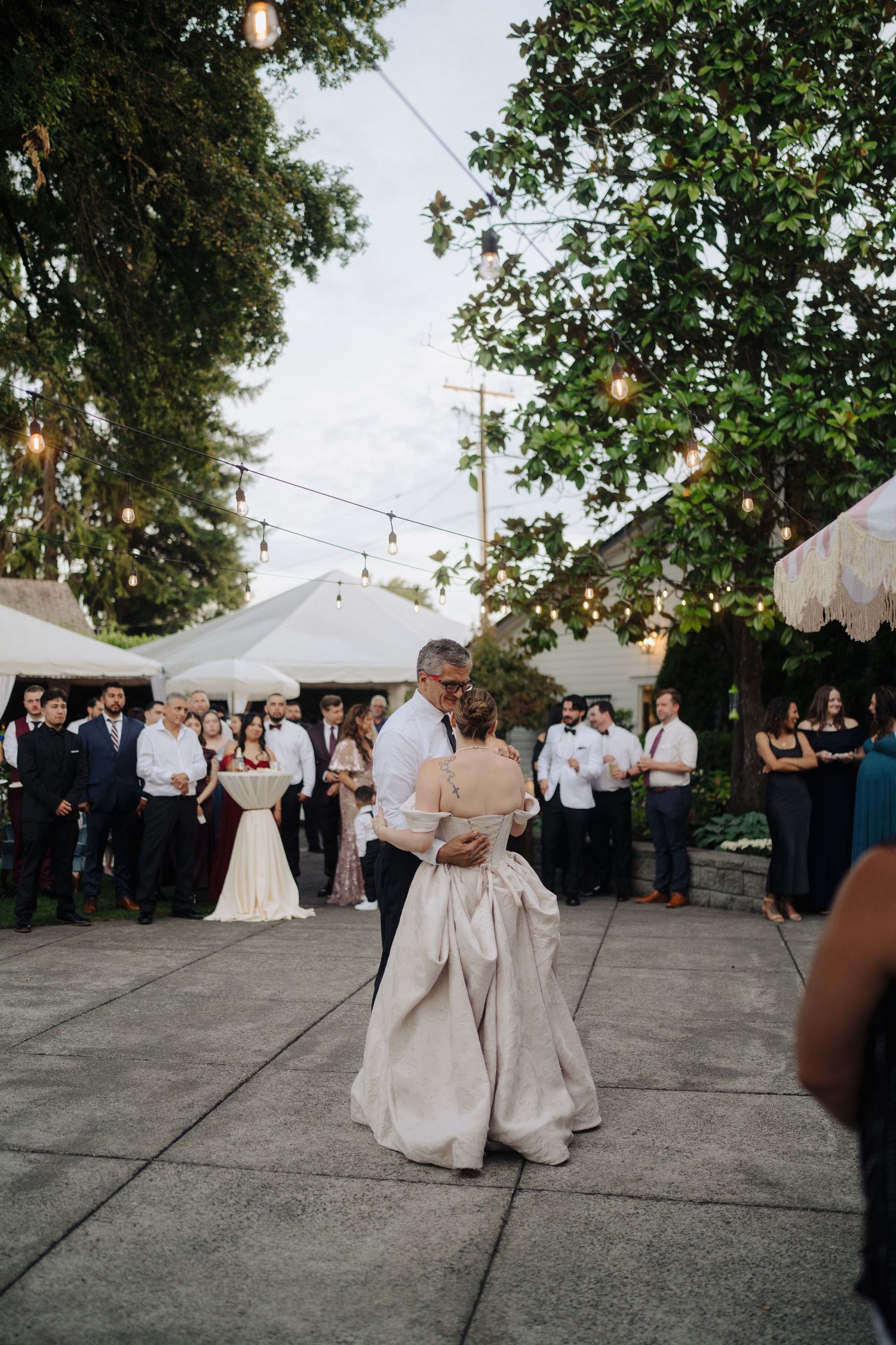 Wedding reception under string lights with guests gathered around outdoor dance floor at dusk.