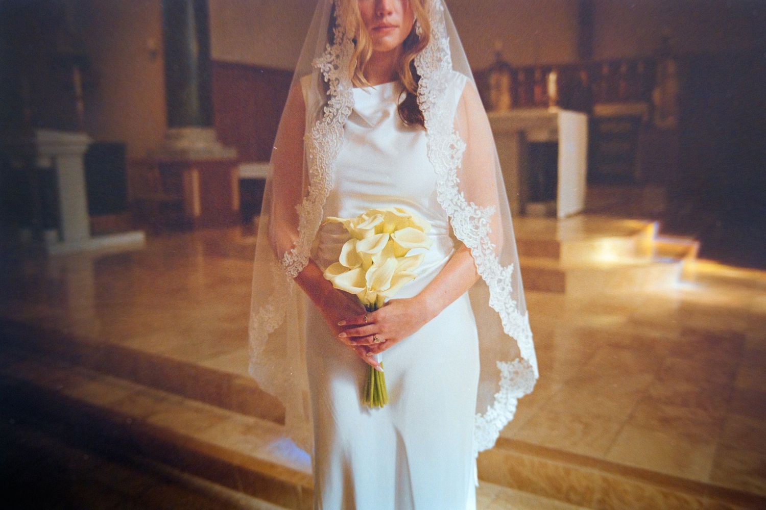 Elegant bride in white dress and lace veil holds calla lily bouquet in a church.