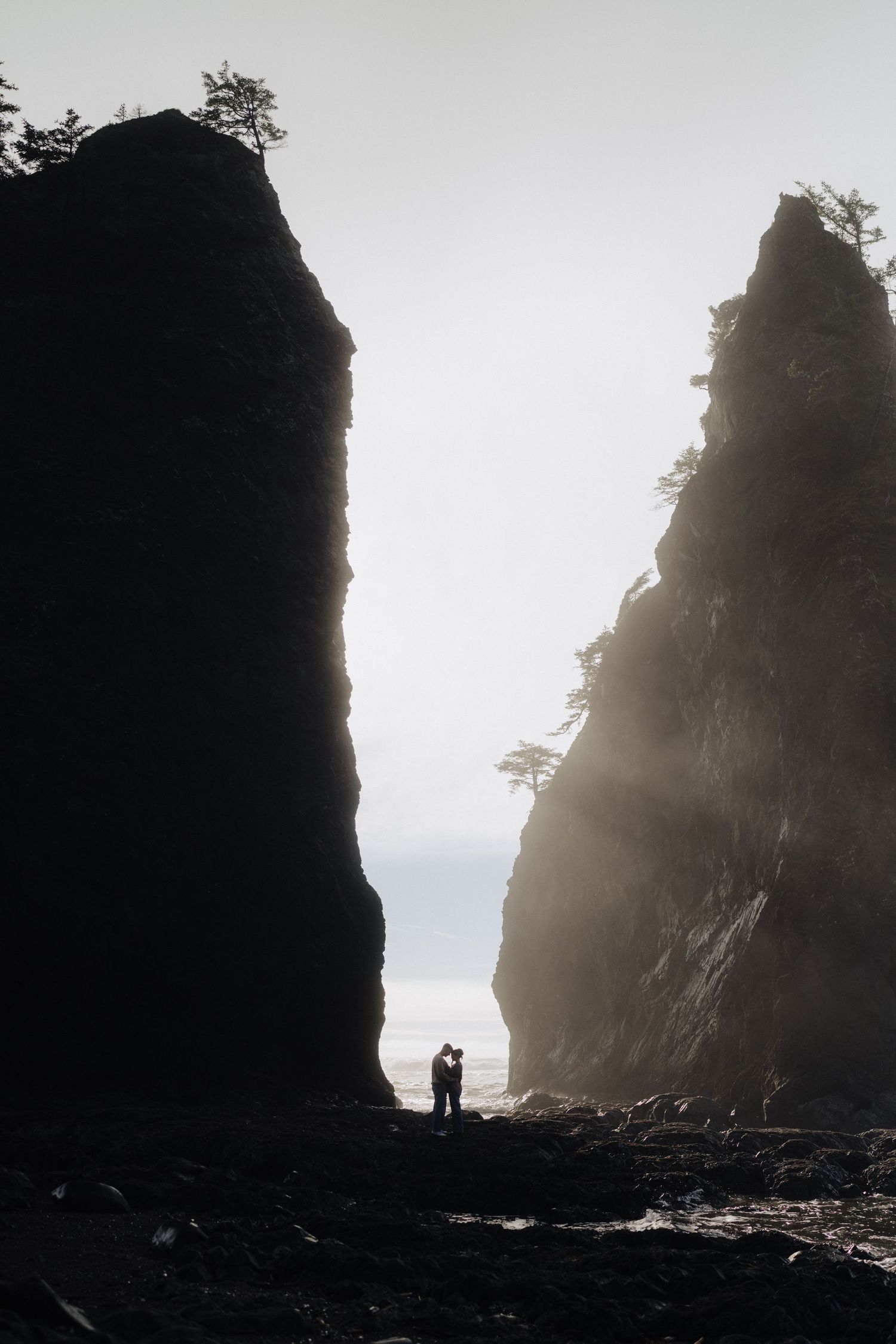 Couple silhouetted between towering sea stacks on a misty Pacific Northwest beach at golden hour.