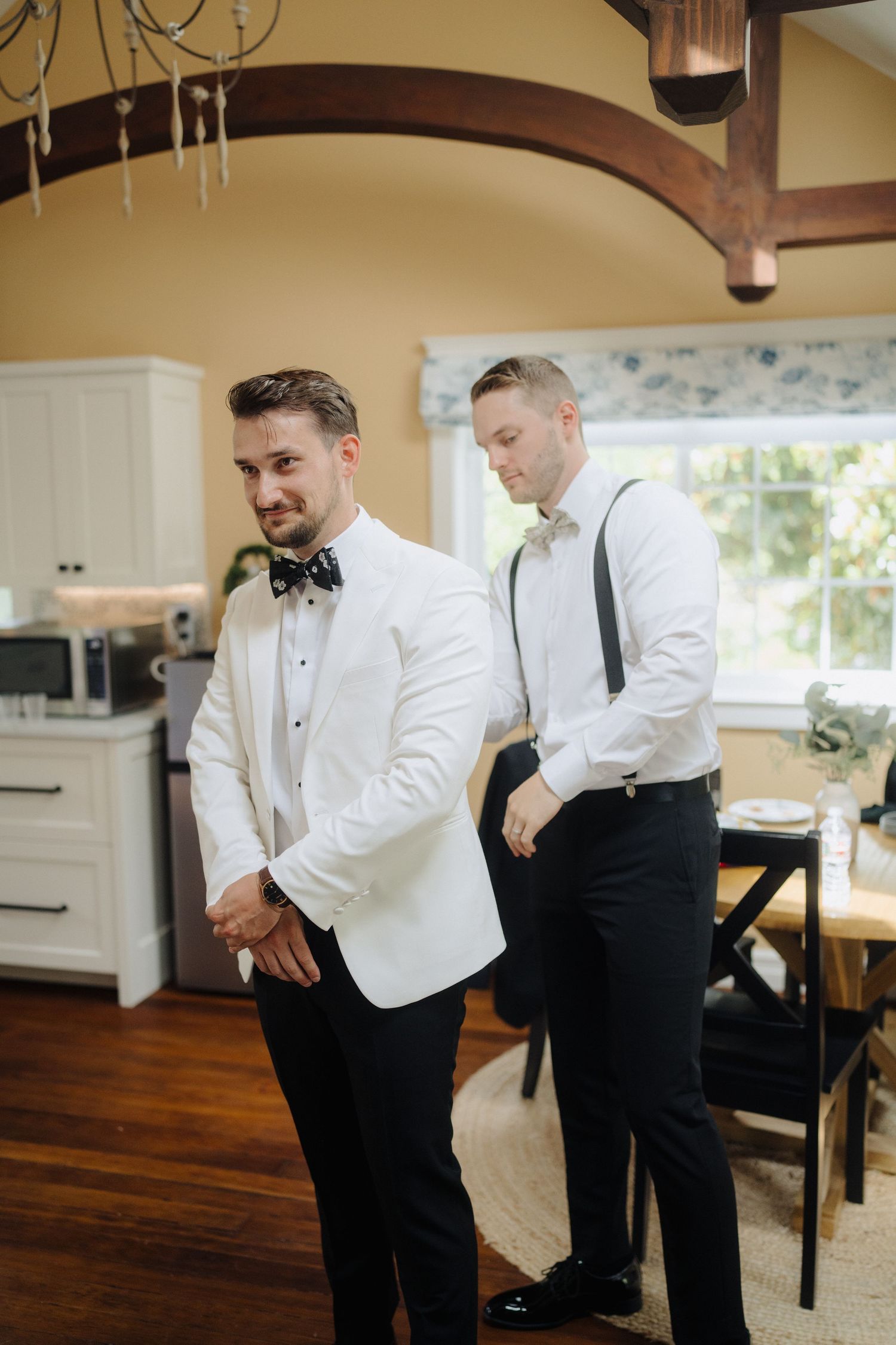 Two groomsmen in white tuxedo jackets and black pants adjusting their bowties before a wedding ceremony.