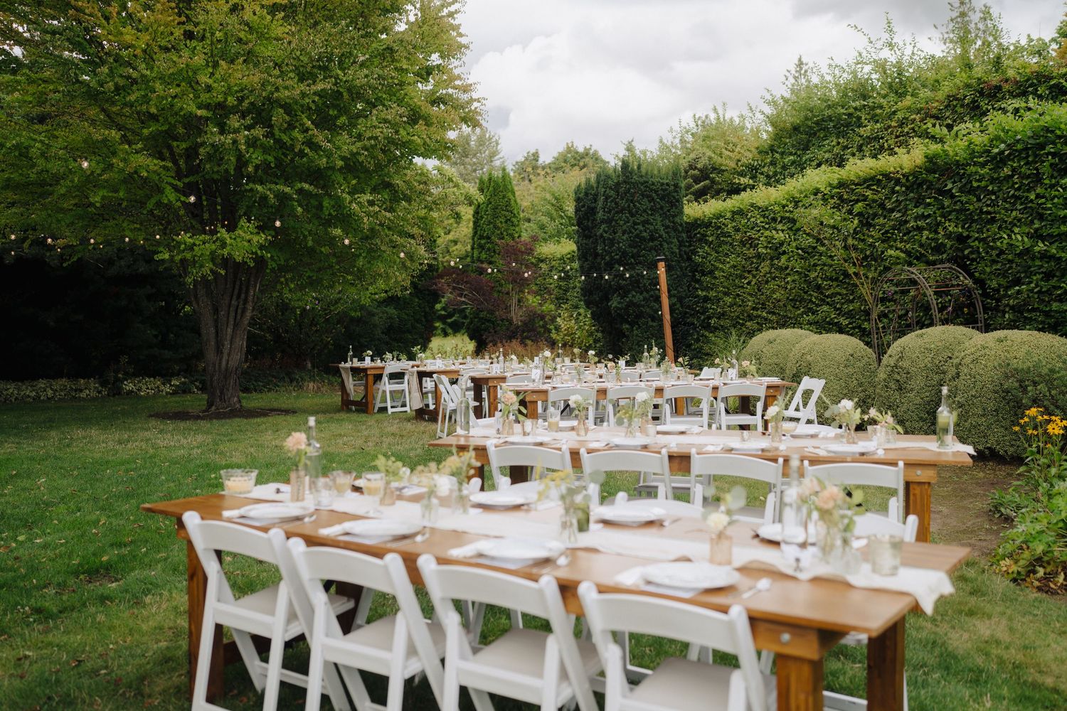 An elegant outdoor wedding reception setup with rustic wooden tables and white place settings in a garden.