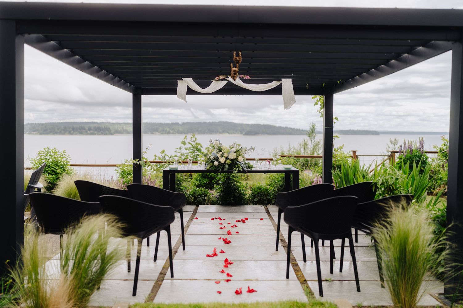 Modern black chairs line an outdoor wedding aisle decorated with red rose petals.