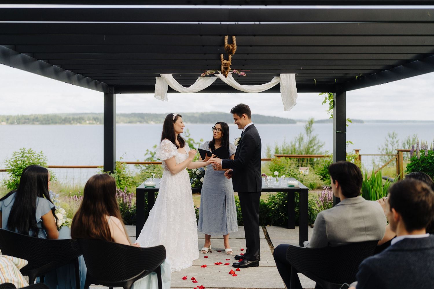An intimate outdoor wedding ceremony takes place under a covered patio overlooking a lake.