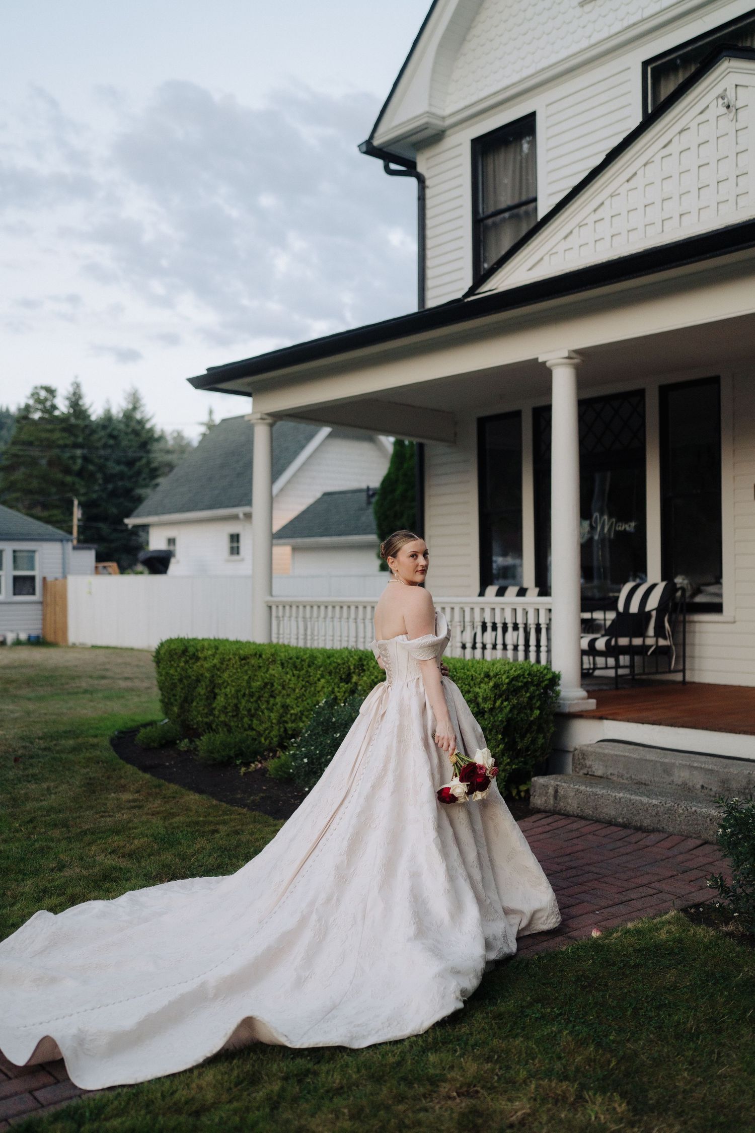 A stunning wedding dress with a dramatic cathedral train flows across a white house's front lawn.