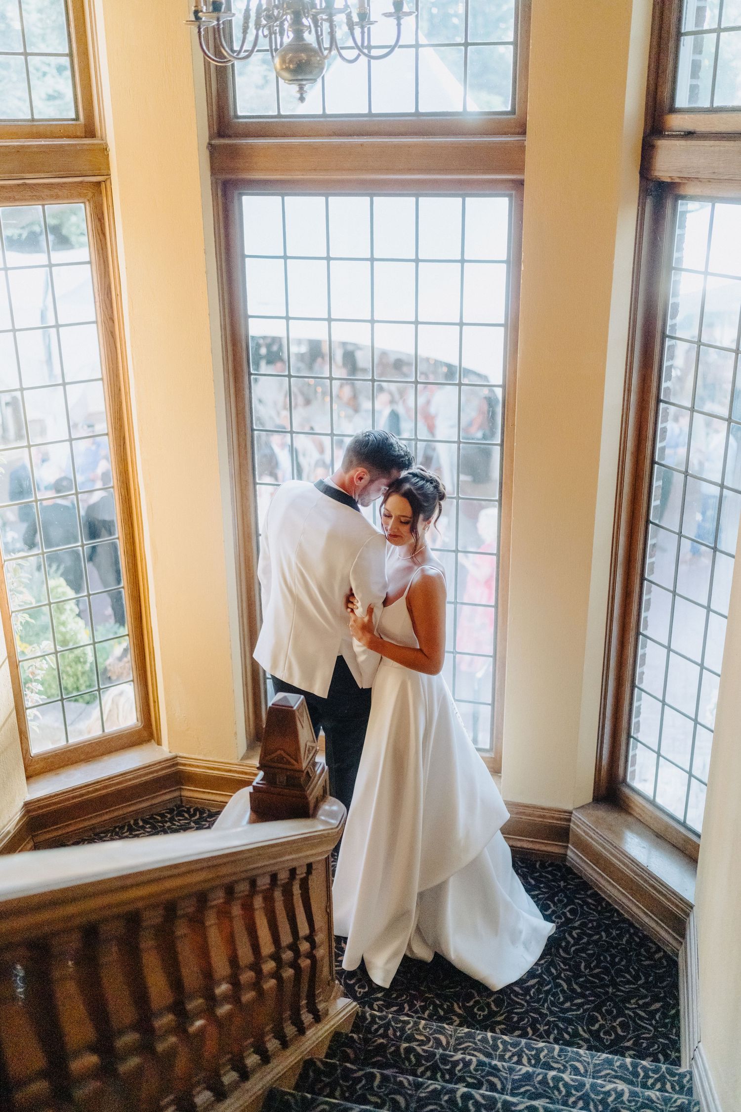 A romantic wedding moment between a couple standing in an elegant window-lit stairwell with patterned carpet.