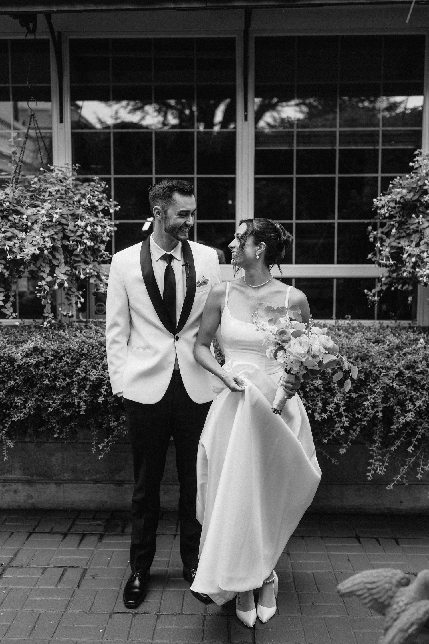 Couple shares playful moment on outdoor patio with flowering bushes in background.