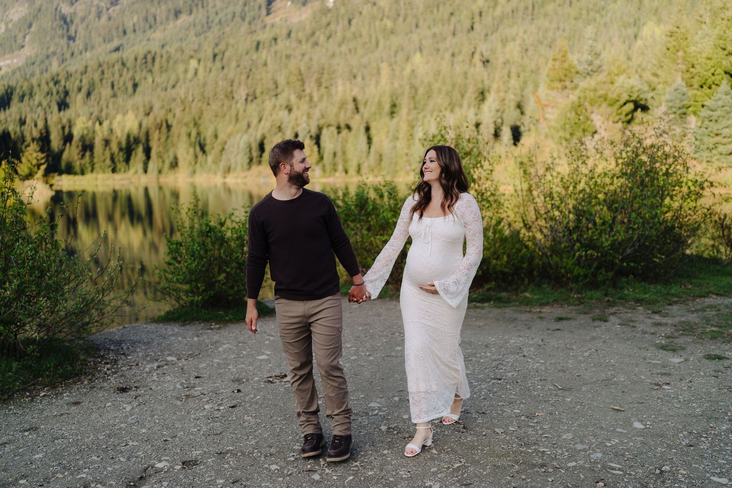 A couple walks hand in hand along a lakeside path with mountains and forest in the background.