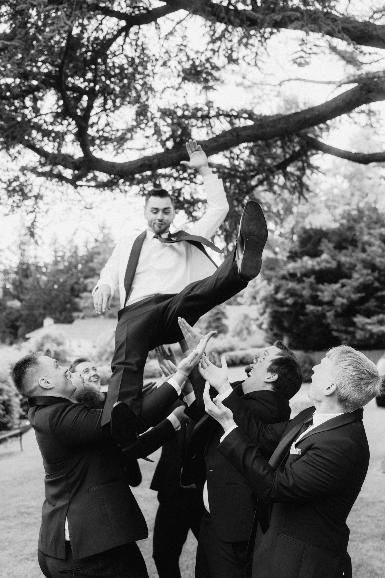 Groomsmen in black suits drinking and celebrating together outdoors under a tree.
