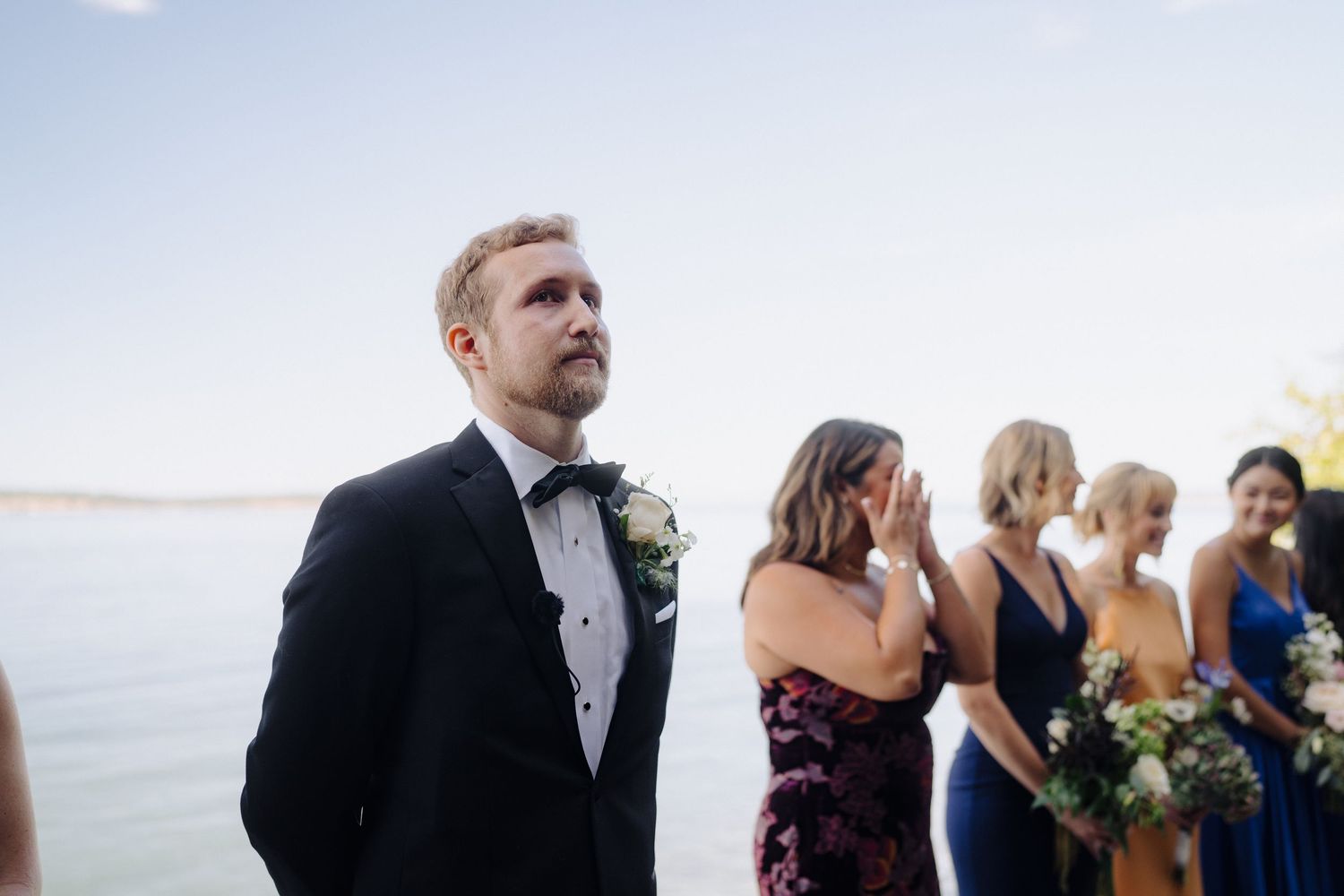 Wedding ceremony guests stand together outdoors with an ocean view on a bright sunny day.