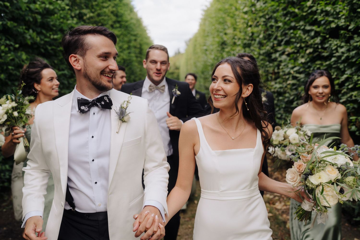 Wedding party members in white and formal attire celebrate joyfully in a garden setting.