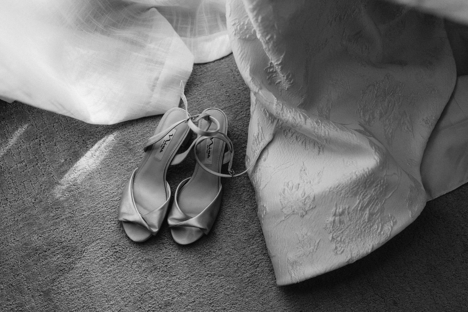 Black and white photo of a bride's wedding dress train and shoes trailing behind her on a textured floor.