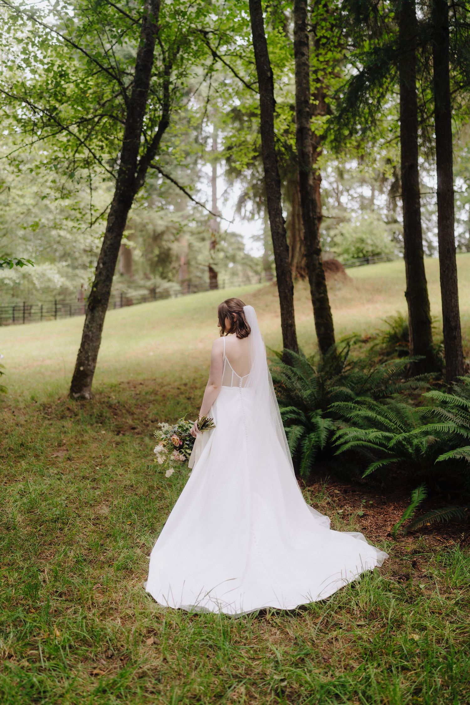A bride in a flowing white dress and veil stands among trees and ferns in a serene woodland setting.